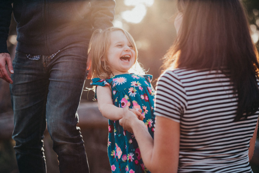 A little girl lets out a huge laugh while holding her mama's hands