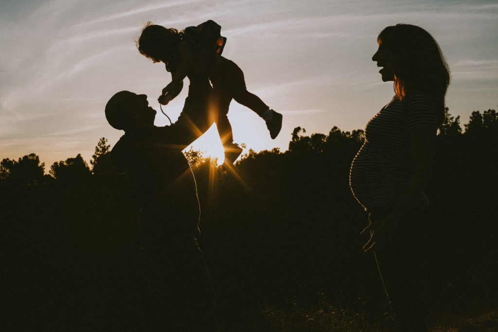 A silhouette family portrait - dad lights daughter high in the air while pregnant mom watches at sunset