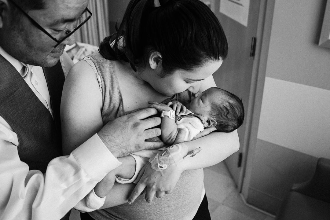 Mom Kissing Newborn Baby In Hospital While Father Looks On Black and White Photo